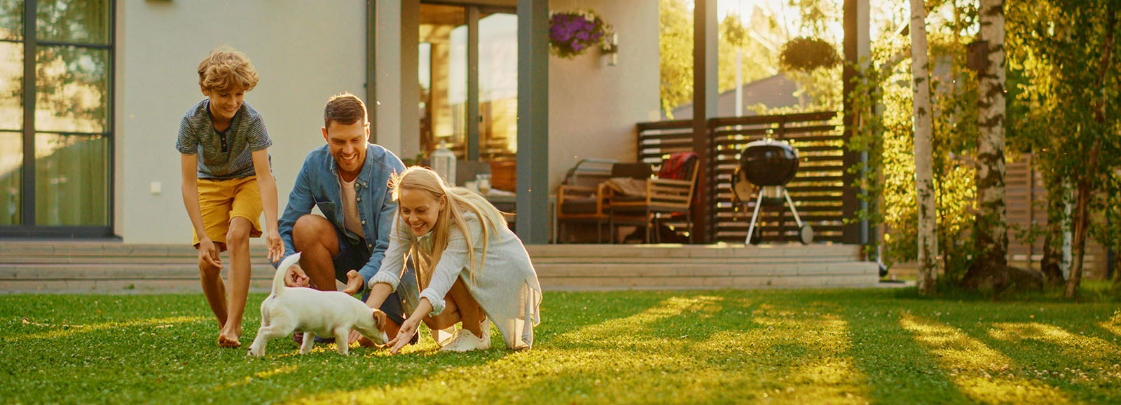 Smiling Father, Mother and Son Pet and Play with Smooth Fox Terrier Retriever Dog. Sun Shines on Idyllic Happy Family with Loyal Pedigree Dog have Fun at the Idyllic Suburban House Backyard